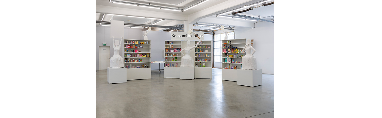 In der Halle unten der Kunsthalle Göppingen stehen mehrere weiße Skulpturen auf Podesten. Die Skulpturen stellen menschliche Figuren dar und sind minimalistisch gehalten. Im Hintergrund befinden sich mehrere weiße Regale, die mit bunten Verpackungen und Konsumgütern gefüllt sind. Über den Regalen steht das Wort 