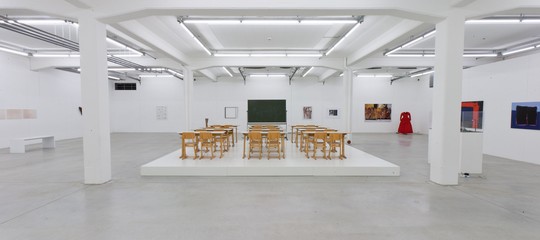 Das Bild zeigt einen großen, weißen Ausstellungsraum, die Halle oben in der Kunsthalle Göppingen, mit moderner Beleuchtung. In der Mitte des Raumes steht ein Podest mit alten Holzschulbänken und einer Tafel, die an ein Klassenzimmer erinnert. An den Wänden hängen verschiedene Kunstwerke, darunter Gemälde und Fotografien. Die Szene wirkt wie eine Mischung aus Klassenzimmer und Kunstausstellung.