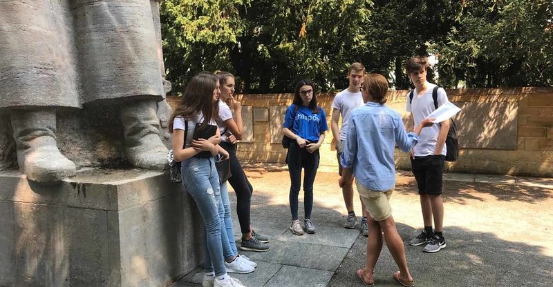 Eine Gruppe Jugendlicher steht im Freien neben einem großen Steindenkmal. Eine Frau mit einem Zettel in der Hand spricht zur Gruppe, die aufmerksam zuhört. Im Hintergrund sind Bäume und eine Mauer zu sehen. Beim Klicken auf das Bild startet ein YouTube Video zu diesem Projekt.