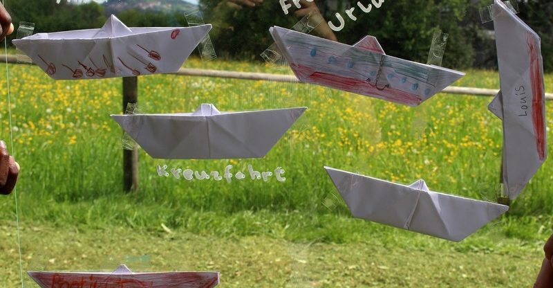 Das Bild zeigt mehrere aus Papier gefaltete und teilweise bemalte Boote, die an einer durchsichtigen Fläche (vermutlich Glas oder Folie) befestigt sind. Dahinter ist eine grüne Wiese mit gelben Blumen zu sehen. Auf den Booten stehen Wörter wie „Kreuzfahrt“ oder Namen wie „Louis“.