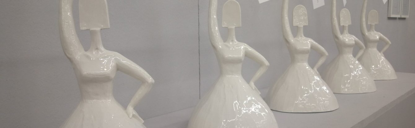 Reihe von weißen, minimalistischen Keramik-Skulpturen in Form von Frauenfiguren mit ausgestrecktem Arm. Die Figuren tragen lange Kleider und haben ovale, abstrahierte Köpfe ohne Gesichtszüge. Die Oberflächen der Skulpturen sind glänzend. Alle Skulpturen stehen in gleicher Pose, mit einer Hand in die Hüfte gestützt und der anderen erhoben.