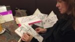 Zu sehen ist eine Frau, die an einem Tisch sitzt und auf ein Blatt Papier zeichnet. Der Tisch an dem sie sitzt ist zugestellt mit weiter Zeichnungen und anderen Dingen und neben ihr auf dem Stuhl liegen auch noch mehr Zeichnungen.