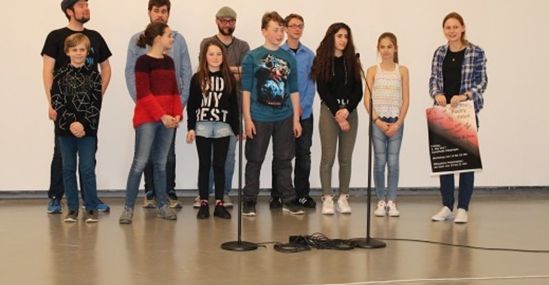 Auf dem Bild steht eine Gruppe von Jugendlichen in der sechsten Klasse und Erwachsene hinter ihnen in einem Raum vor Mikrofonen. Eine Person hält ein Plakat und alle schauen nach vorne und wirken bereit für eine Präsentation oder Aufführung.