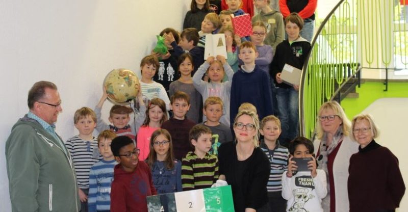 Das Bild zeigt eine große Gruppe von Grundschulkindern, die gemeinsam mit mehreren Erwachsenen auf einer Treppe stehen. Einige Kinder halten Gegenstände wie einen Globus, Bücher oder Schilder in der Hand. Im Vordergrund stehen Erwachsene und Kinder um ein farbiges Würfelmodell mit Zahlen. Die Stimmung wirkt fröhlich und lebendig.