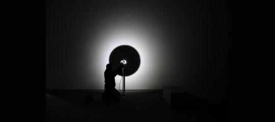 Silhouette einer Person, die vor einer Wand mit einem starken Lichtkegel kniet. Die Person hält eine Lampe, deren Licht einen kreisförmigen Schatten auf die Wand wirft. Der Rest des Bildes ist dunkel, wodurch das Licht und die Silhouette im Zentrum hervorgehoben werden.