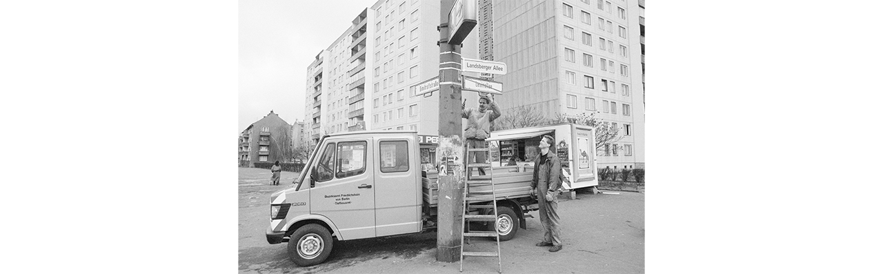 Daniel Biskup, die Welle der Umbenennung, Berlin-Ost, 1991, dargestellt wird ein LKW hinter einem Betonpfsoten mit Straßenschildern, zwei Männer in Arbeitskleidung, ein Mann auf einer Trittleiter am Montieren