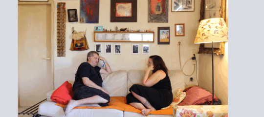In einem gemütlichen Wohnzimmer sitzen ein Mann und eine Frau einander gegenüber auf einem Sofa. Beide sind entspannt, barfuß und in ein Gespräch vertieft. Die Wände sind mit zahlreichen Bildern, Fotos und kleinen Dekorationsgegenständen geschmückt, über dem Sofa hängen gerahmte Kunstwerke und eine kleine Ablage mit Figuren und Fotos. Auf dem Sofa liegen bunte Kissen und eine Decke, daneben steht eine Stehlampe mit floralem Lampenschirm, die eine warme Atmosphäre erzeugt.