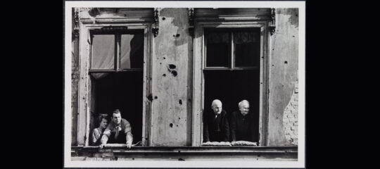 Fotografie zweier Fenster an einer heruntergekommenen Hausfassade. In dem linken Fenster stehen zwei junge Männer, einer von Beiden lehnt sich etwas aus dem Fenster heraus, während der Andere sich eher im Hintergrund hält. Im rechten Fenster stehen zwei alte Männer, sie lehnen sich etwas heraus. Alle vier Männer schauen in die selbe Richtung, es scheint, als würden etwas beobachten.